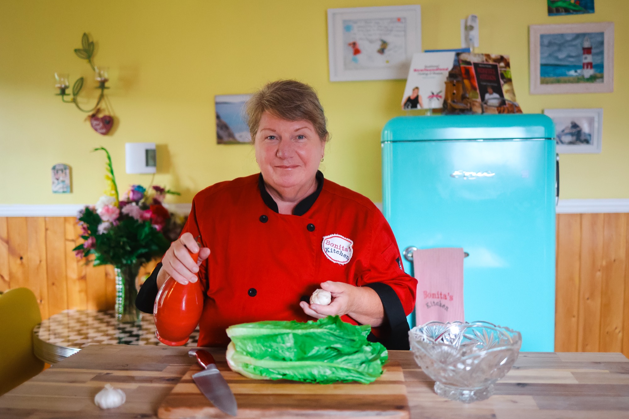 Bonita in her kitchen, in her red chef's jacket, with the turquoise fridge behind her